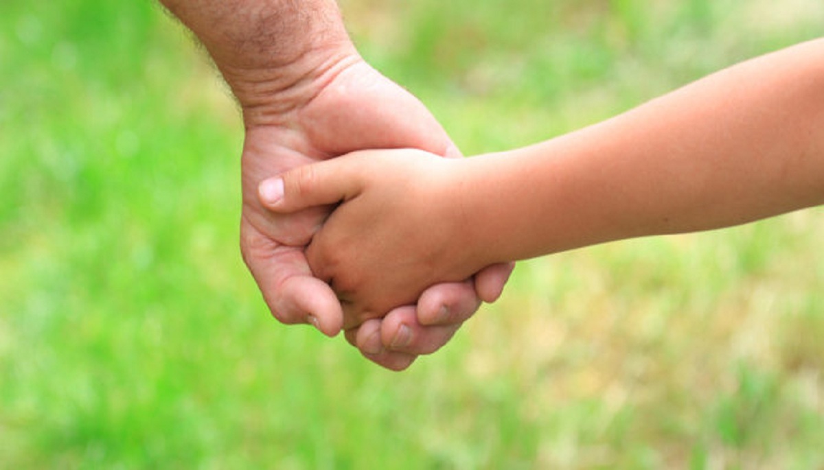 a father's hand holds a child's hand against a background of green grass