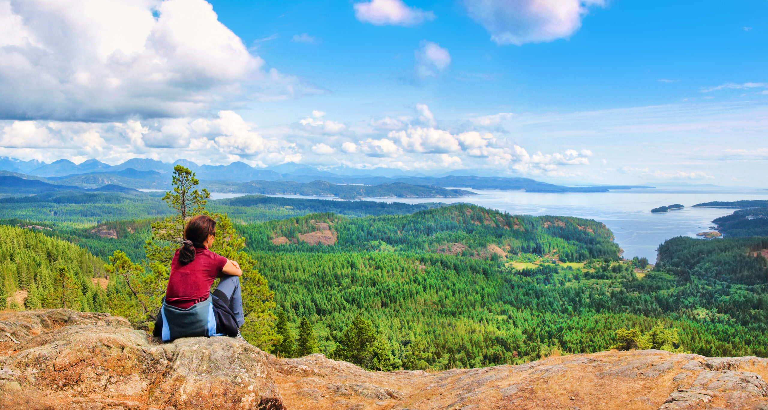 woman sits at the top of a mountain and looks down on the forest and ocean scenery on Vancouver Island, British Columbia