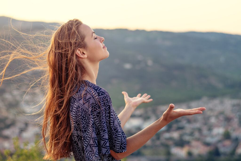 young woman with long hair stands on a hill overlooking the city, eyes closed, in silent worship and giving thanks to God