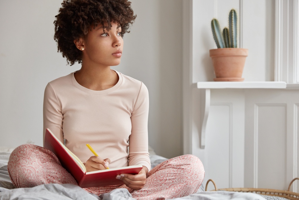 Journaling for Anxiety Relief - a thoughtful Black woman sits on her bed writing in journal - Shutterstock