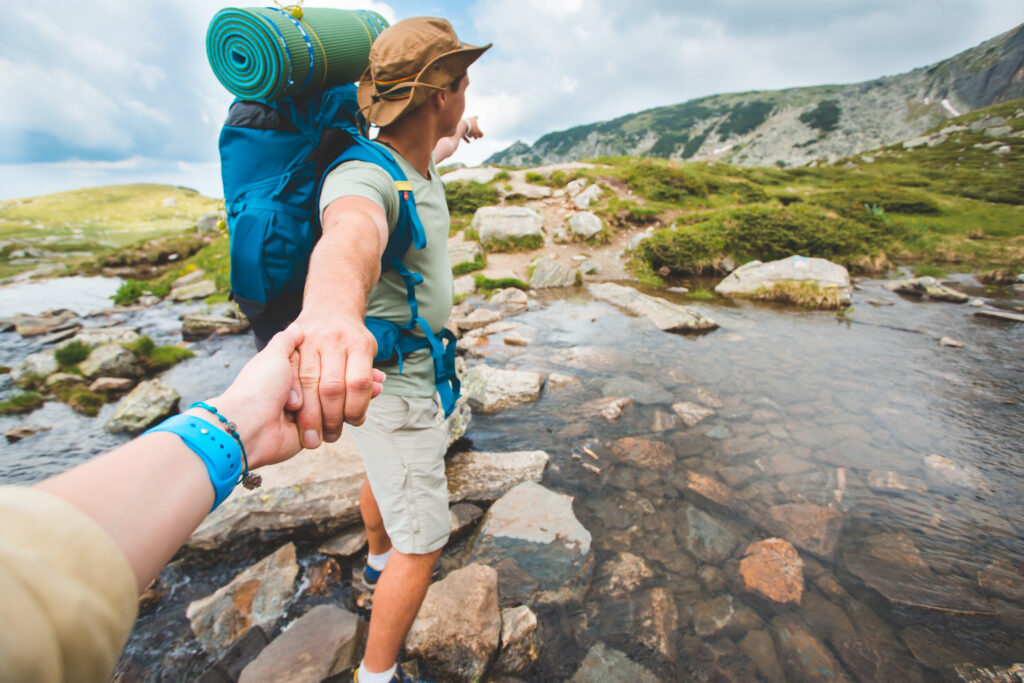 man leads person by the hand crossing a river pointing towards a forest